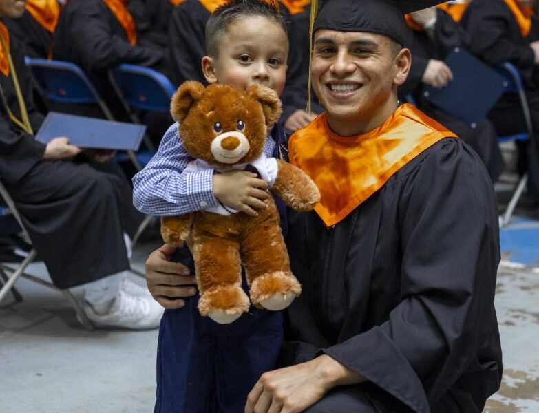 Graduate in cap and gown smiling and posing with a young boy holding a stuffed teddy bear