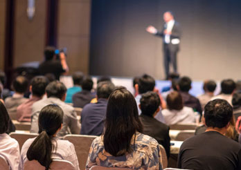 Audience seated and listening to a speaker presenting on stage at a conference or seminar.