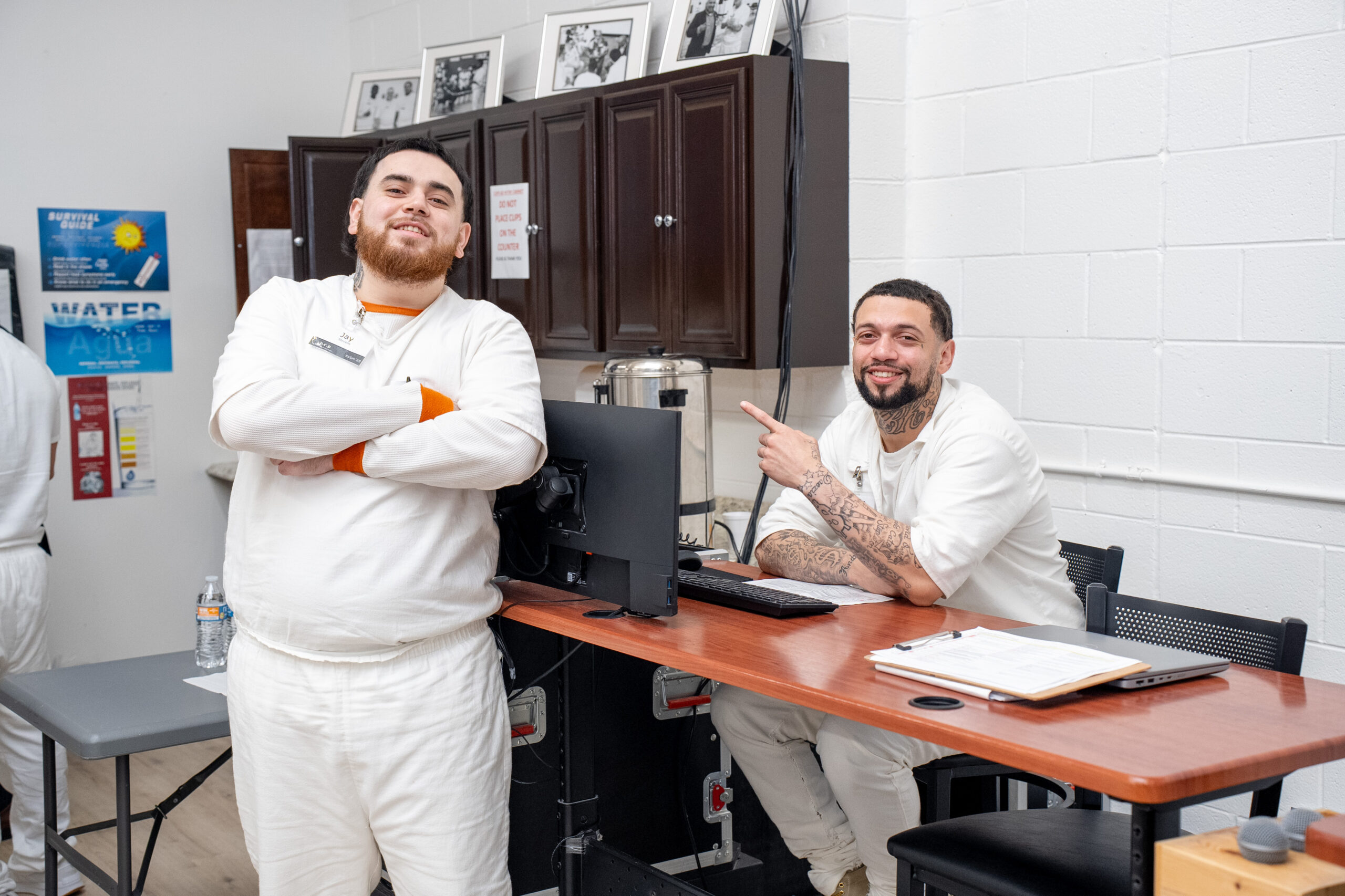 Two PEP participants share a laugh while working behind the scenes, one pointing playfully as they manage event operations from a computer station