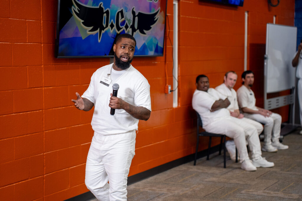 Prison Entrepreneurship Program participant delivers a business pitch with microphone as peers look on during a workshop in a Texas prison
