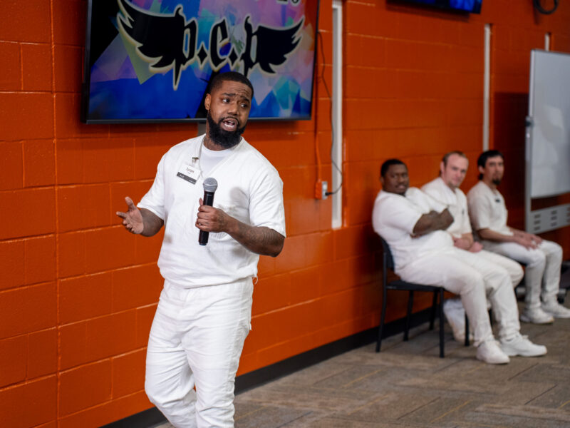 Prison Entrepreneurship Program participant delivers a business pitch with microphone as peers look on during a workshop in a Texas prison