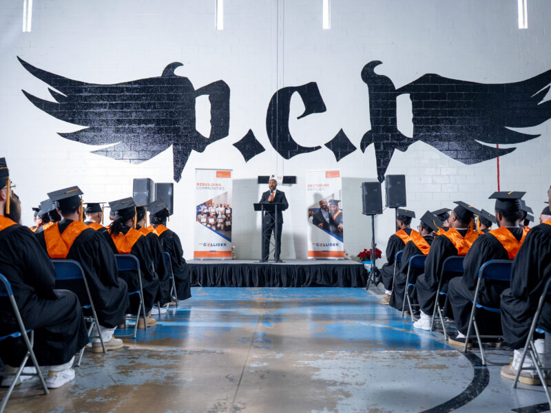 Graduates in caps and gowns attend a Prison Entrepreneurship Program ceremony at a Texas correctional facility, with speaker at podium and PEP logo on wall
