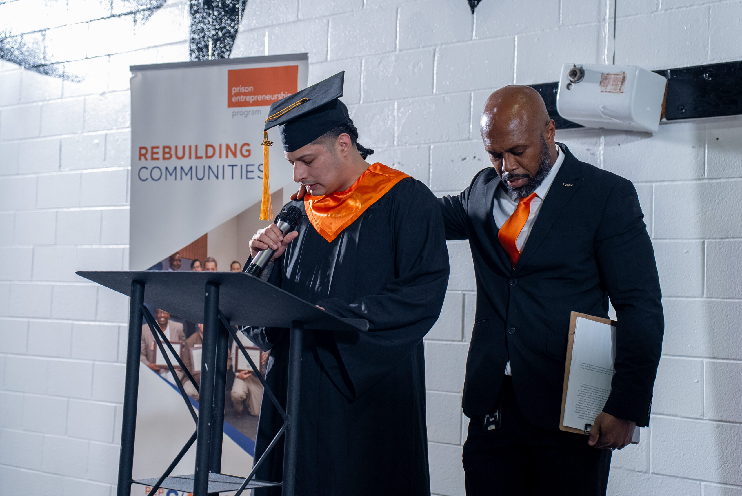 Prison Entrepreneurship Program graduate speaks at the podium during a cap and gown ceremony, with a mentor standing supportively beside him