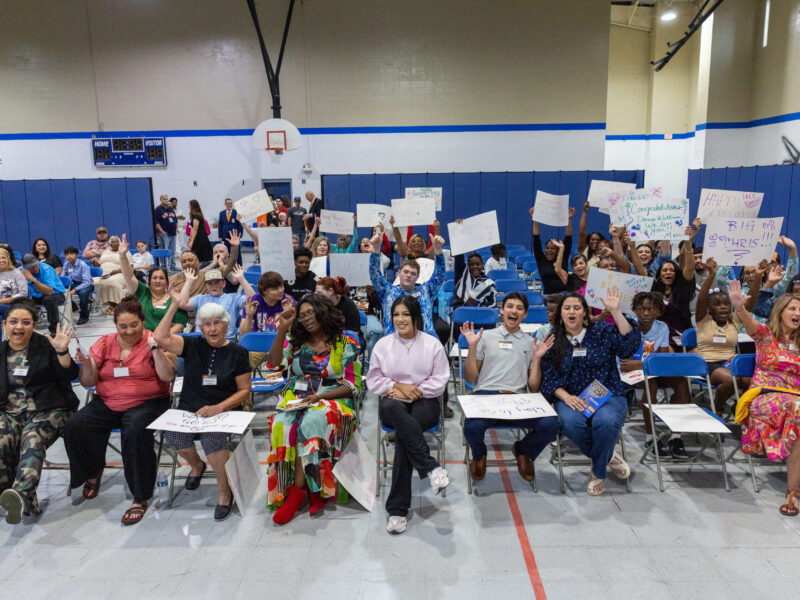 Excited family members and supporters holding handmade signs and cheering at a PEP graduation event inside a gymnasium
