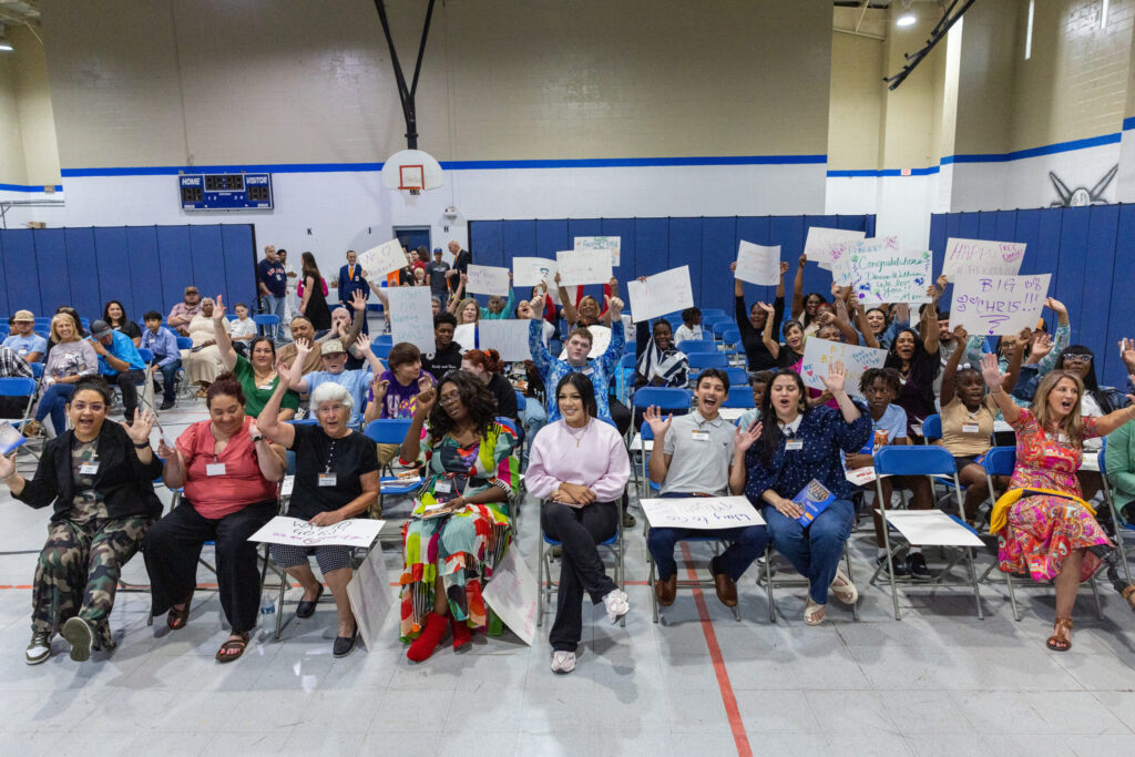 Excited family members and supporters holding handmade signs and cheering at a PEP graduation event inside a gymnasium