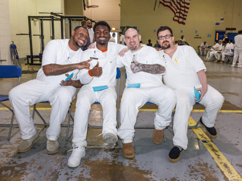Incarcerated men participating in the Prison Entrepreneurship Program smile and give thumbs up during a PEP class session in a Texas prison gym