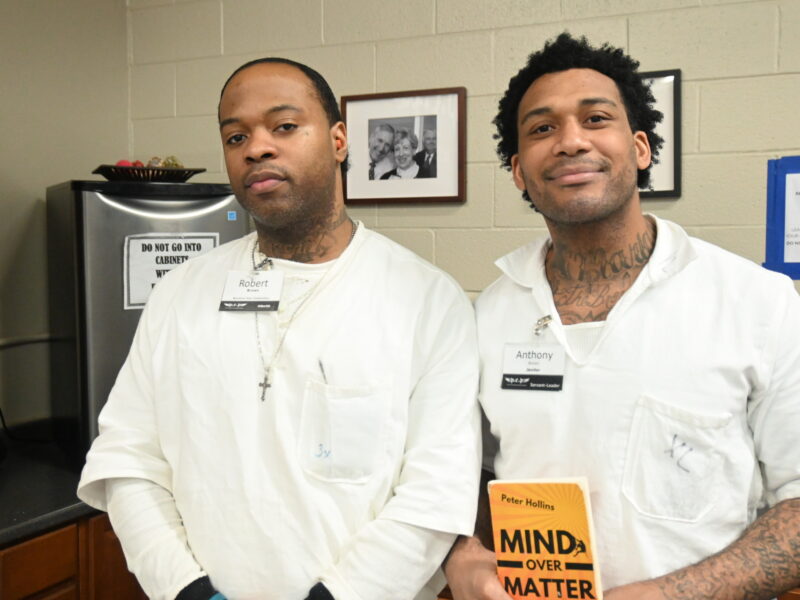 Two PEP participants in white uniforms standing together, one holding a personal development book titled "Mind Over Matter" by Peter Hollins