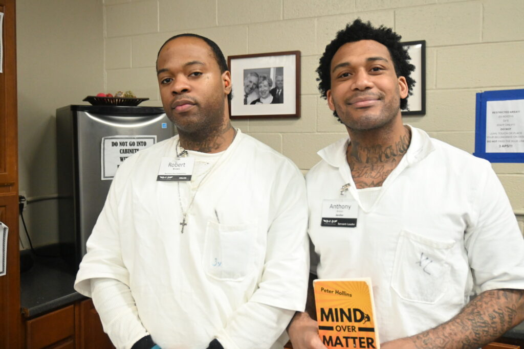 Two PEP participants in white uniforms standing together, one holding a personal development book titled "Mind Over Matter" by Peter Hollins