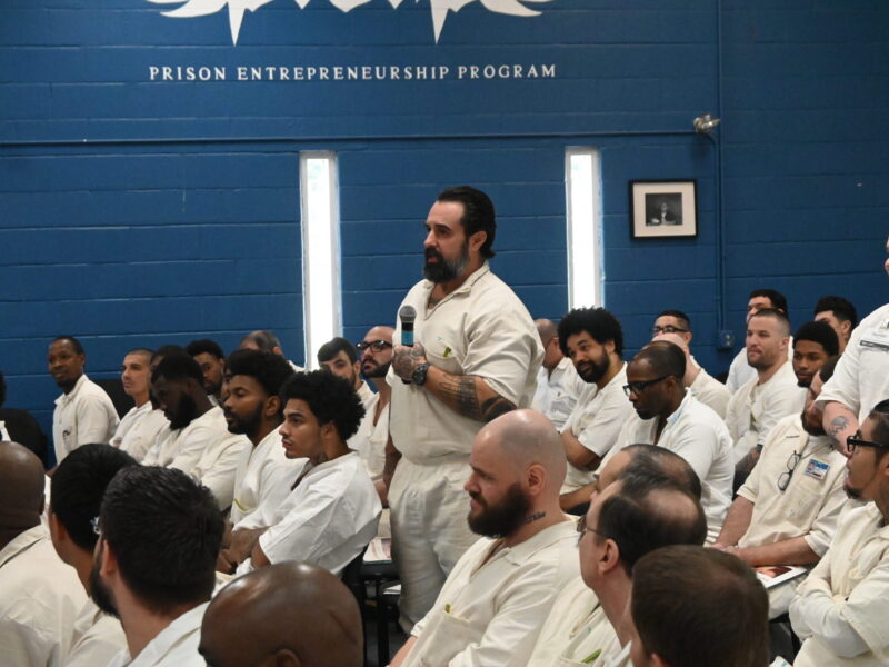 Incarcerated man participating in a Prison Entrepreneurship Program event, standing and speaking into a microphone during a session with fellow PEP participants