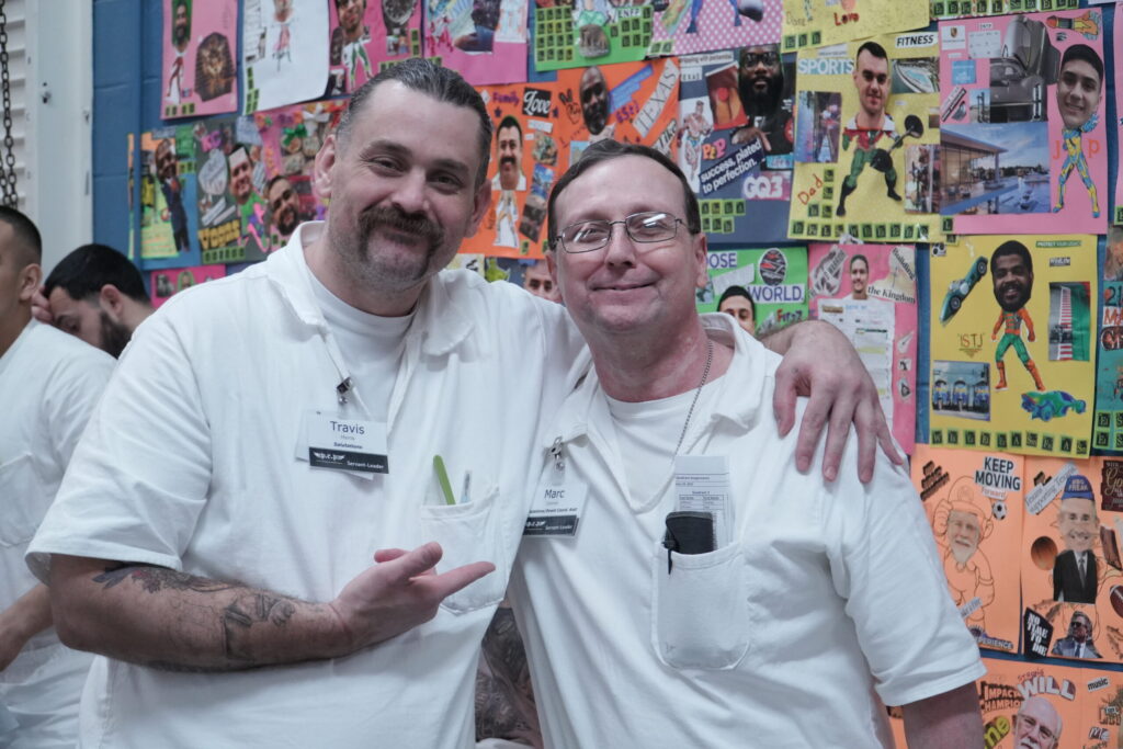 Two PEP participants in white uniforms smiling and posing together in front of colorful vision boards at a Prison Entrepreneurship Program event