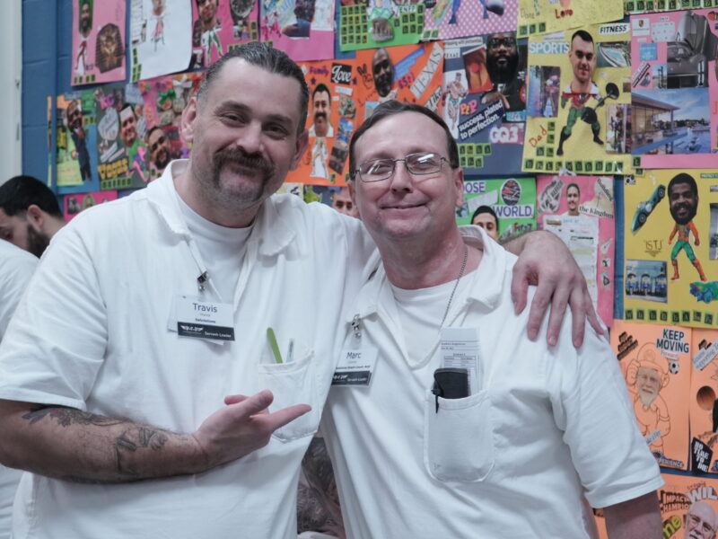 Two PEP participants in white uniforms smiling and posing together in front of colorful vision boards at a Prison Entrepreneurship Program event