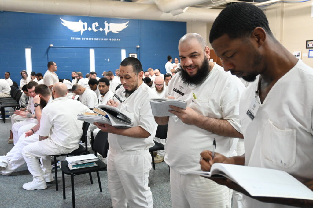 PEP participants in white uniforms engaged in reading and writing during a classroom session under a Prison Entrepreneurship Program banner
