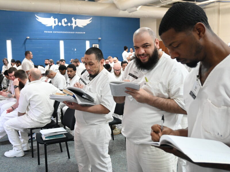 PEP participants in white uniforms engaged in reading and writing during a classroom session under a Prison Entrepreneurship Program banner